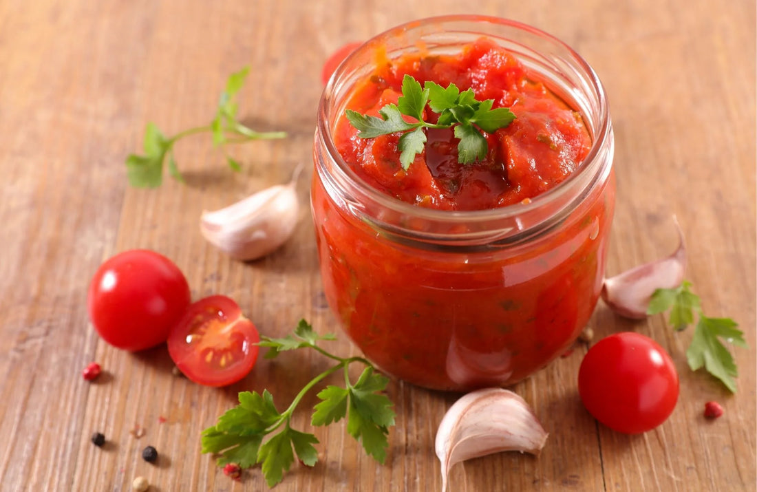 A glass jar filled with a delicious tomato sauce garnished with parsley. Surrounding the jar are whole and halved cherry tomatoes, garlic cloves, and sprigs of parsley on a wooden surface.