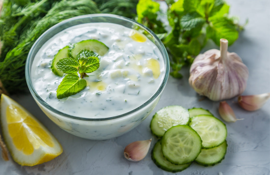 Homemade Greek tzatziki in a white bowl, topped with olive oil and fresh herbs, served with grilled pita bread and cucumber slices – fresh summer dip made with yogurt, garlic, and cucumber.